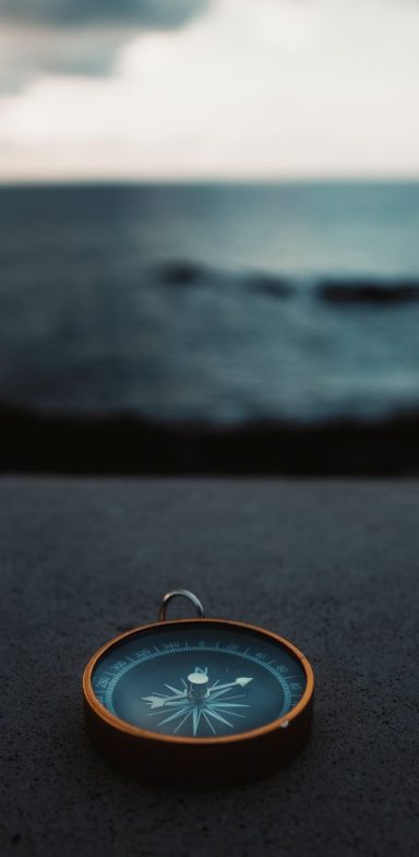 Ein Kompass liegt auf einer Oberfläche, mit unscharfem Hintergrund von Wasser und Himmel.