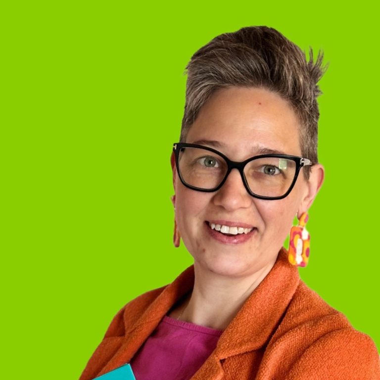 Frau mit Brille, modernen Ohrringen und einem bunten Outfit vor grünem Hintergrund.