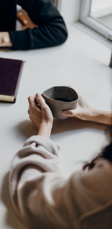 Person hält eine Tasse, während eine andere Person am Tisch sitzt.
